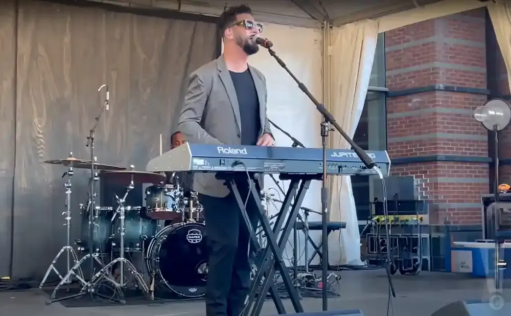 A man in a grey suit jacket sings into a microphone while playing a Roland Jupiter-50 keyboard at an outdoor concert.