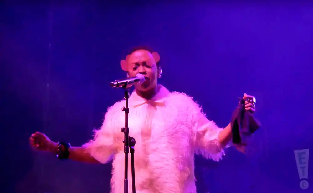 A person in a fluffy white coat and bear ears sings into a microphone on a purple-lit stage.