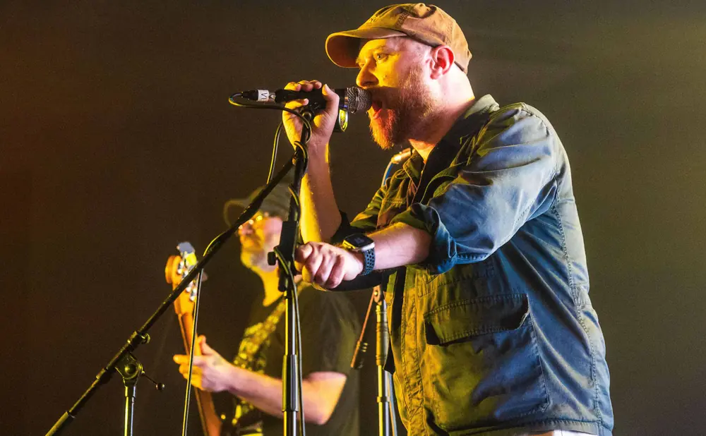 A man with a beard and a brown cap sings into a microphone on stage, with another musician in the background.