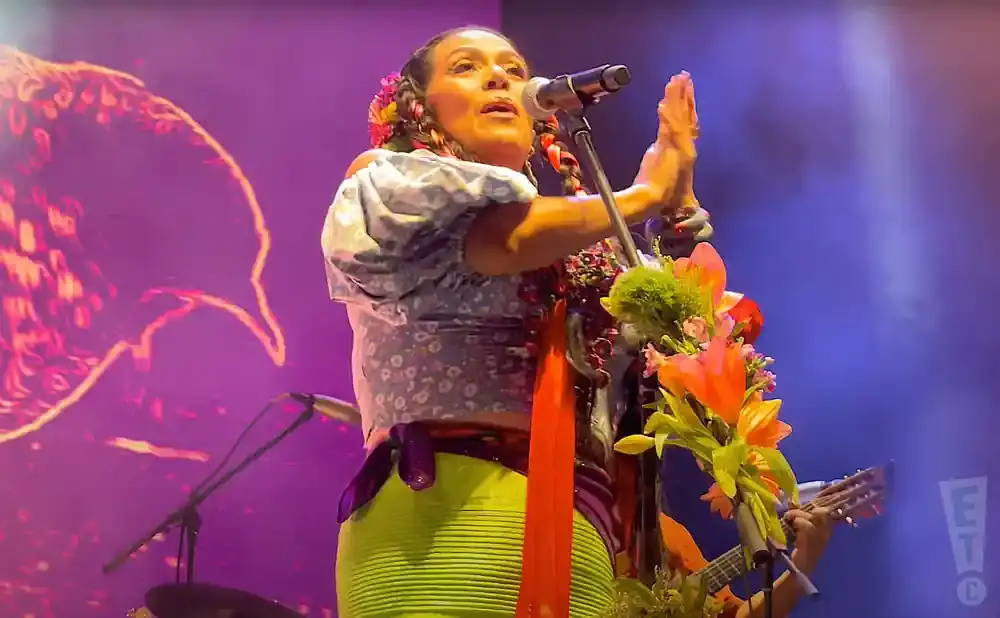 A woman in a floral dress and green skirt sings into a microphone on stage, adorned with flowers.