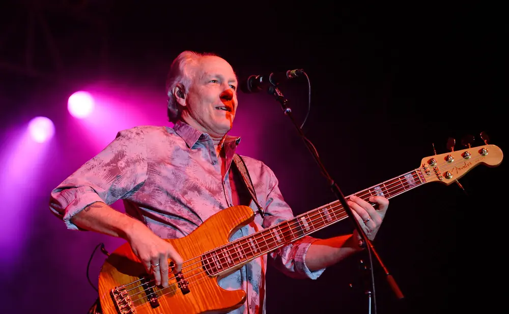 A man plays a Fender bass guitar on stage with purple lighting.