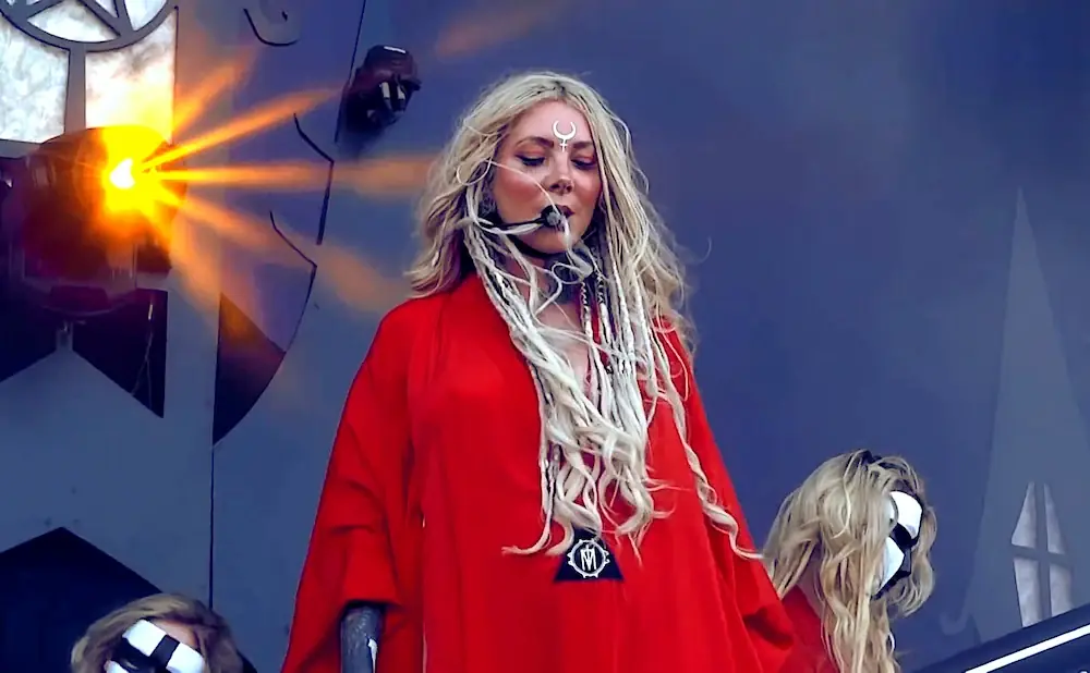 A woman with blonde hair and a red robe sings on stage, with other masked performers around her.