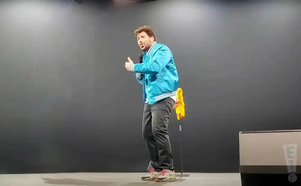 A man in a blue bomber jacket and gray pants holds a microphone, gesturing with his hands while on stage.