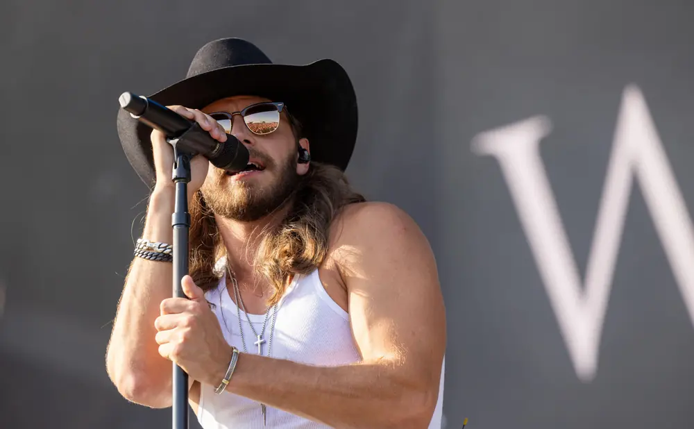 Man in a cowboy hat and white tank top sings into a microphone, wearing sunglasses that reflect a crowd.