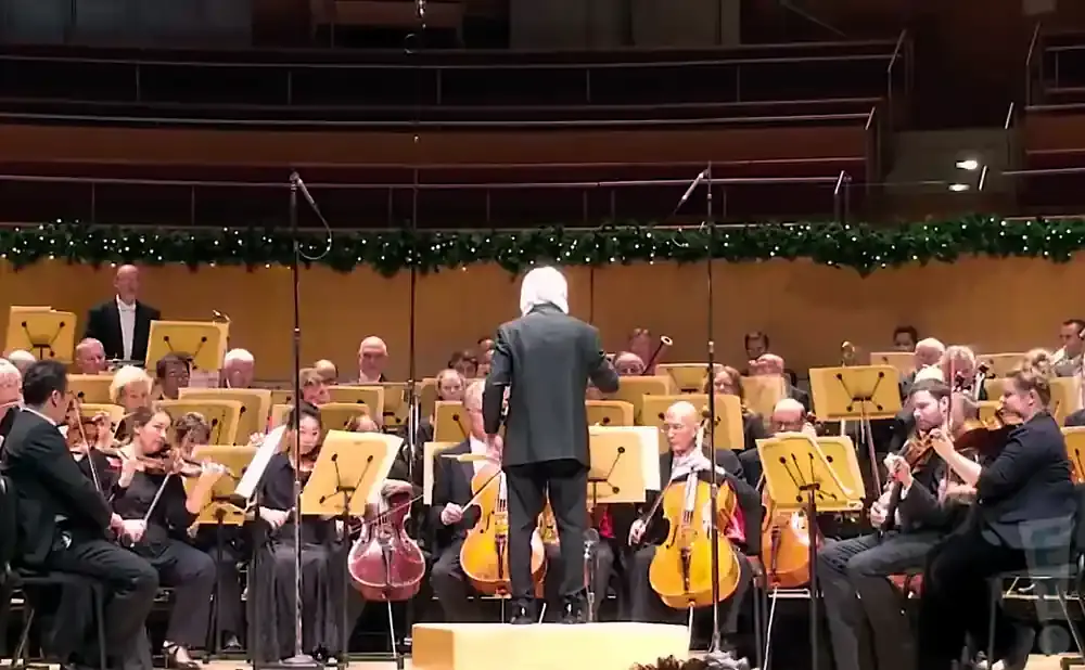 An orchestra performs on stage, led by a conductor with white hair.