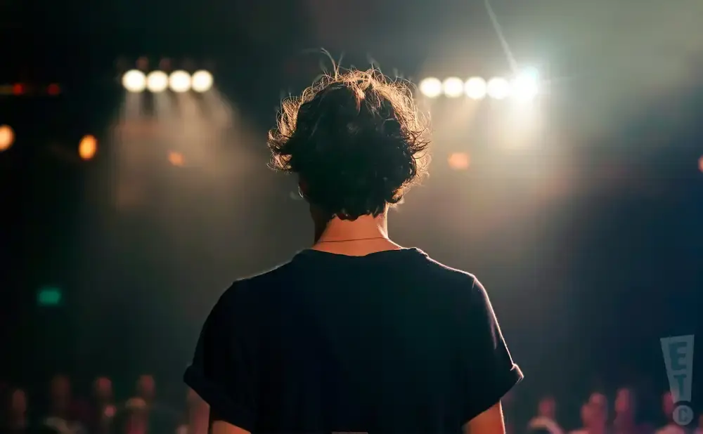 Back of a person with curly hair on a stage, illuminated by stage lights.