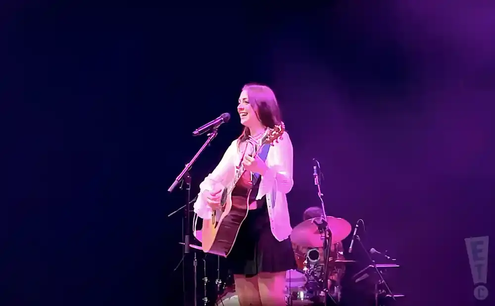 A woman sings and plays an acoustic guitar on stage, bathed in purple light.