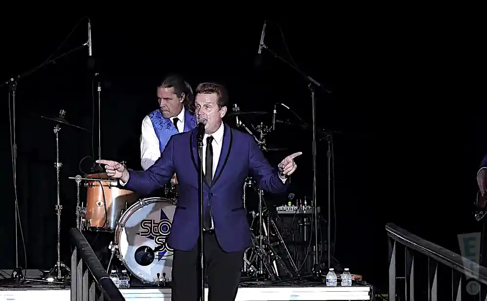 A band performs on stage, with a lead singer in a blue suit at the forefront.