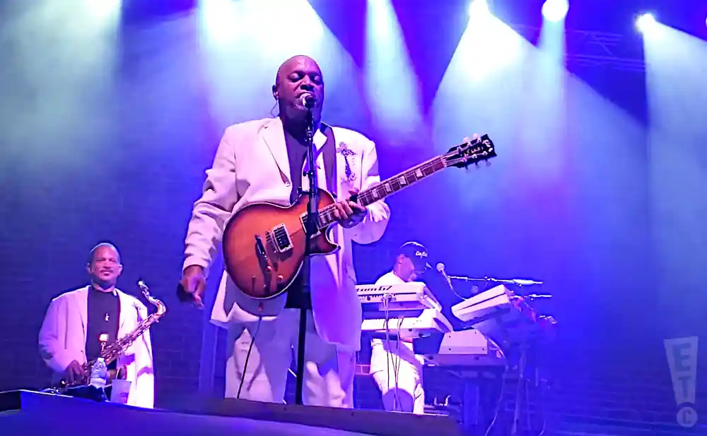 A band performs on stage with a guitarist in a white suit singing, a saxophonist to his left, and a keyboardist behind him.