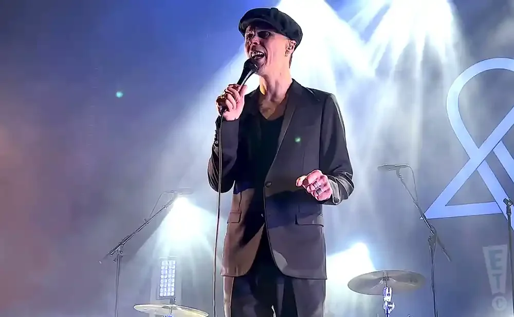 A man in a dark suit and cap sings into a microphone on a stage with bright lights and fog.