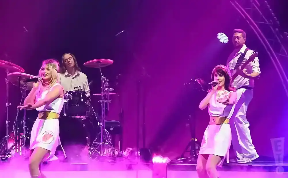 A female singer with blonde hair in a white dress performs on stage with a band.