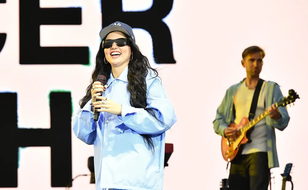 Singer with sunglasses and blue tracksuit performs on stage with guitarist.