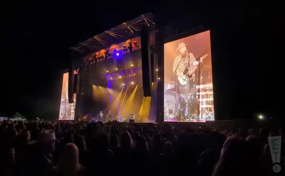 A country singer plays guitar on a large stage with video screens showing his performance to a crowd.