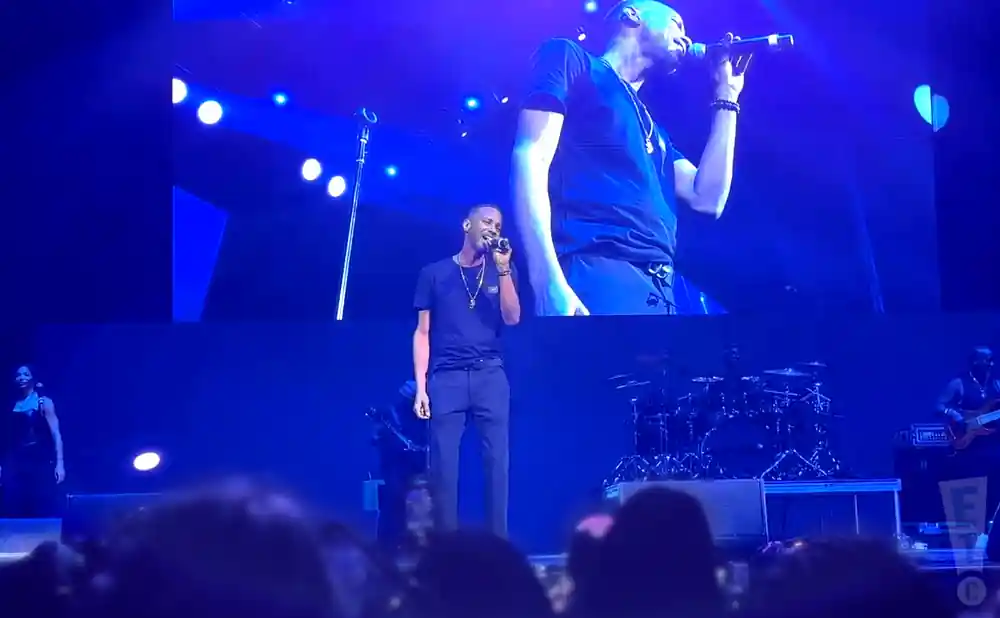 A male singer in a black t-shirt and pants sings into a microphone on a brightly lit stage.