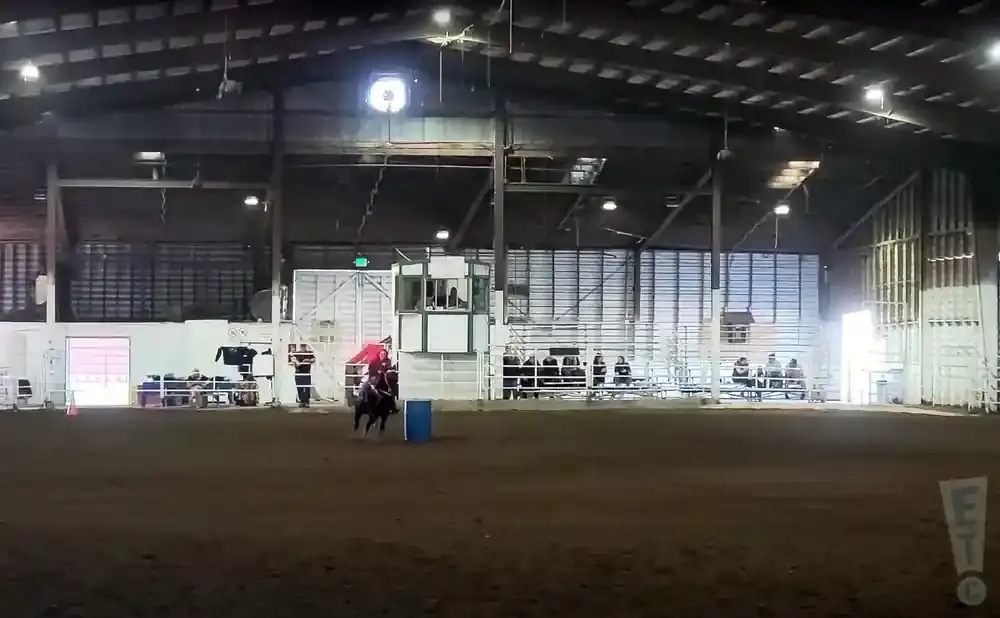 A barrel racer and horse move through an indoor arena, with spectators watching from bleachers.