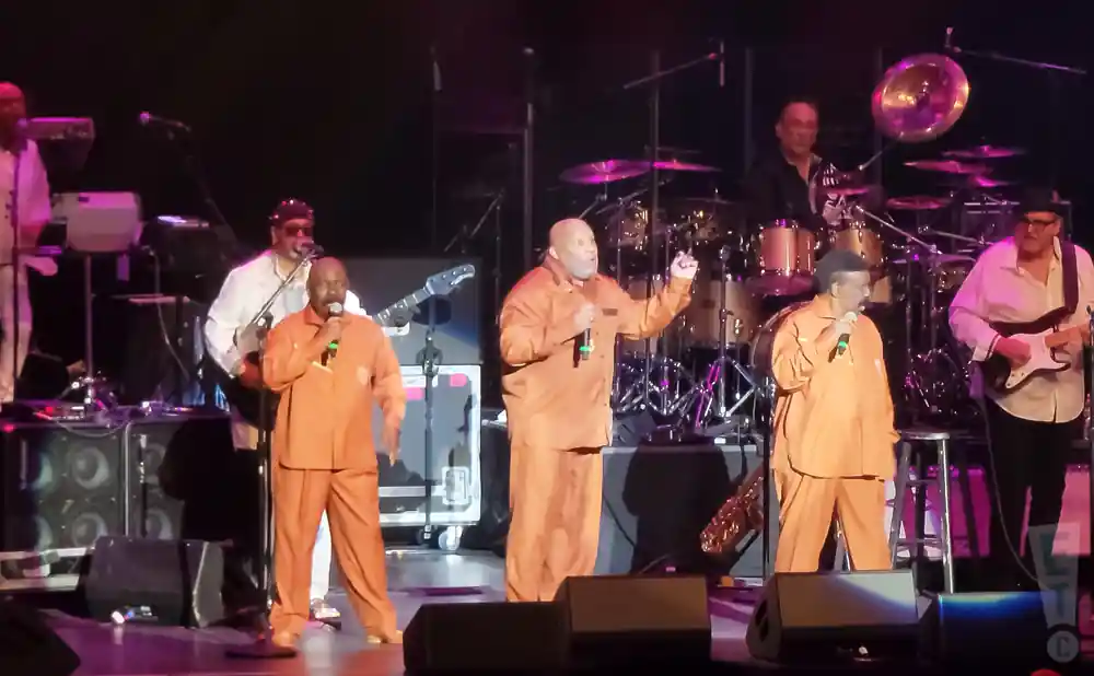 A group of Black men perform on stage in matching orange jumpsuits, singing and playing instruments.