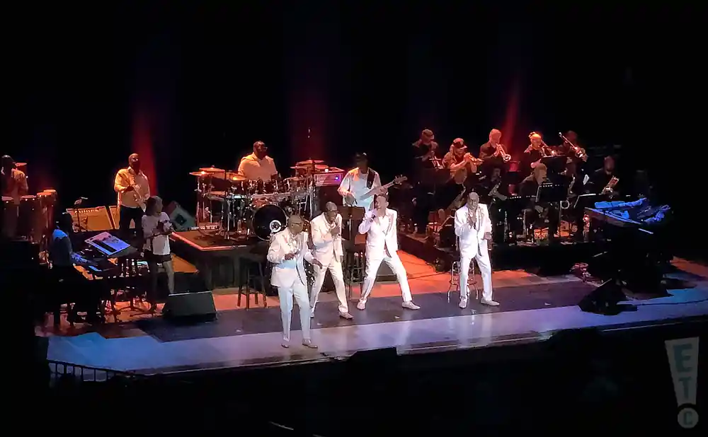 A band performs on stage in white suits, with a horn section and drums in the background.