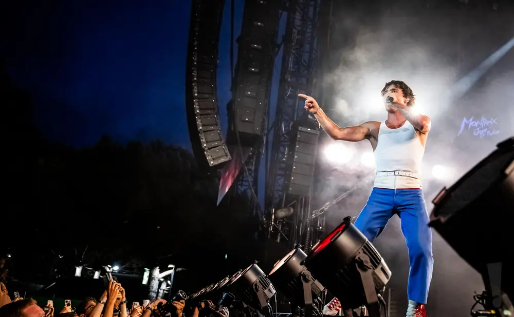 Singer in a white tank top and blue pants points to the crowd at a concert, with stage lights and speakers.