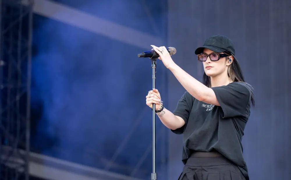 Singer with sunglasses and baseball cap holds microphone on stage.