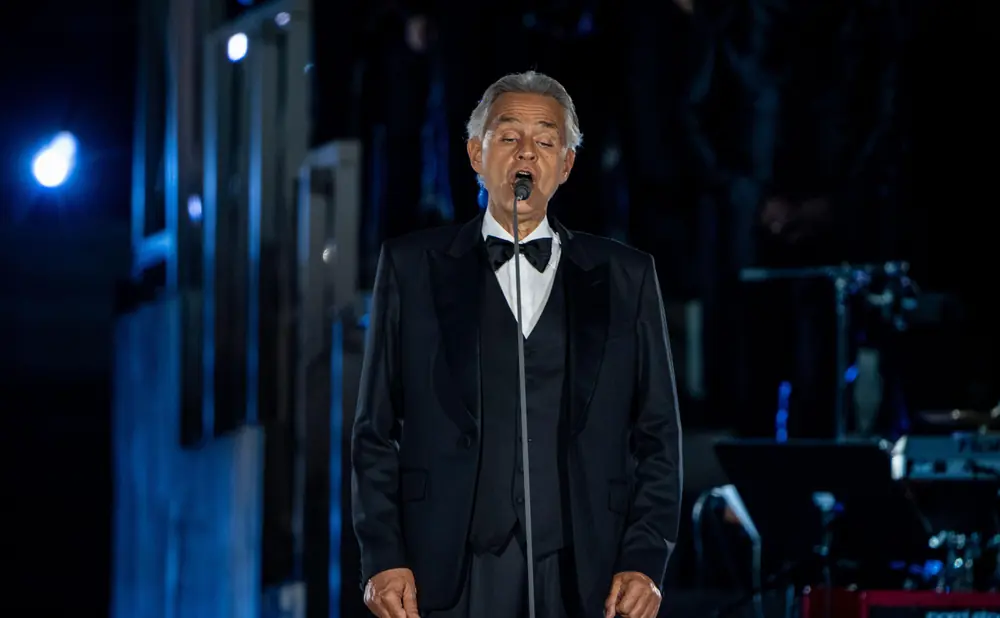 Andrea Bocelli in a tuxedo sings into a microphone on stage.