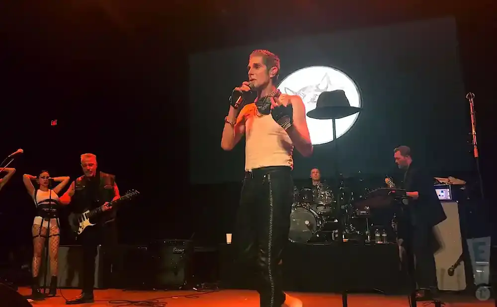 A male singer in a tank top and leather pants performs on stage with a band.