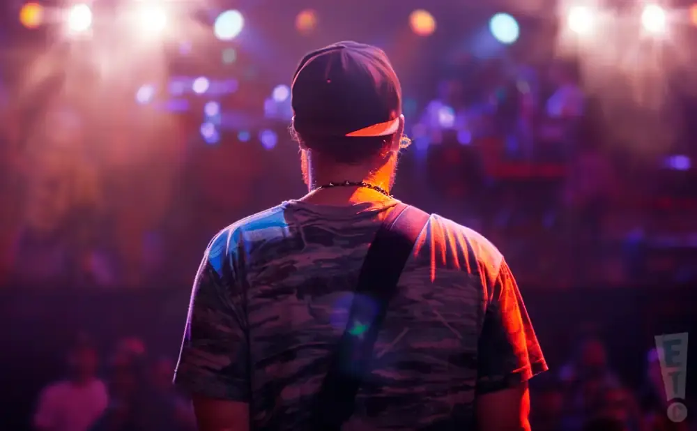 Back view of a person wearing a baseball cap and camouflage shirt on stage, with concert lights and a crowd in the background.