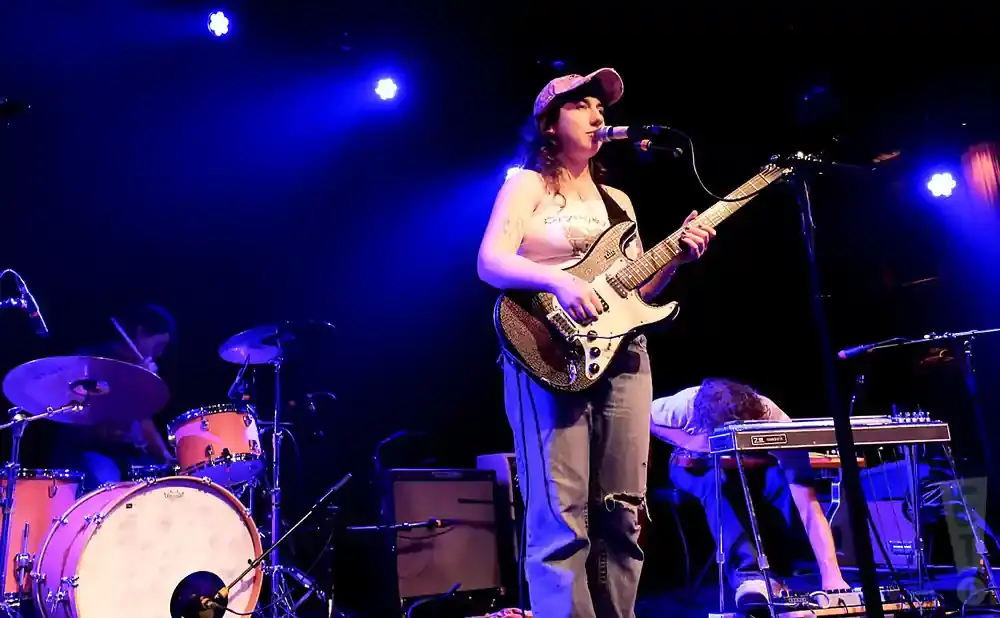 A band performs on a dimly lit stage with blue spotlights. A female guitarist sings into a microphone, while a drummer plays behind her.