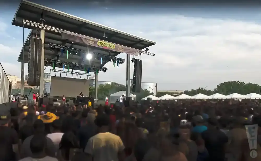Outdoor concert with a large crowd gathered in front of a stage under a partly cloudy sky.