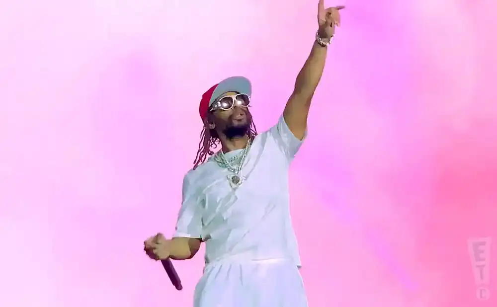 Rapper Offset performs on stage in a white outfit, red and blue hat, and sunglasses, pointing up with a microphone in his hand.