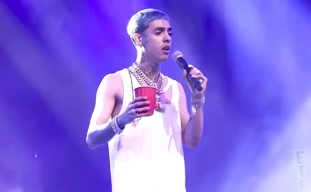 A man with blue hair and tattoos holds a red cup and a microphone, performing on stage under purple lights.