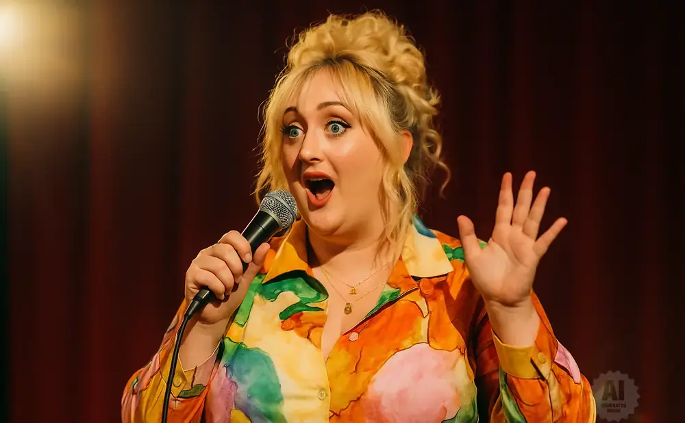 A woman with blonde hair in a messy bun, wearing a colorful shirt, holds a microphone and gestures with her hand while speaking.