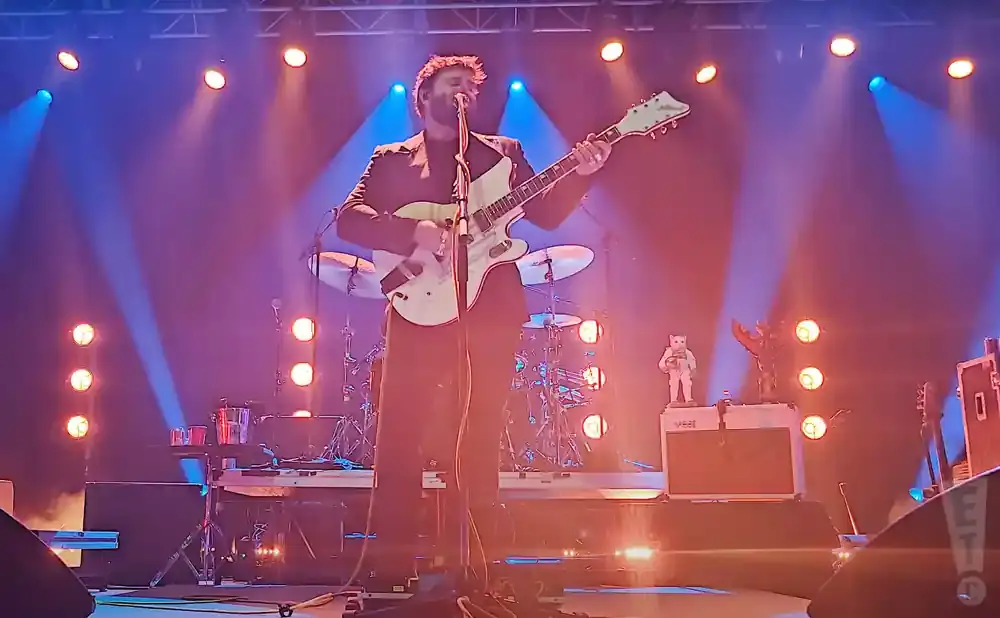A musician plays a white electric guitar on stage, bathed in blue and orange lights.