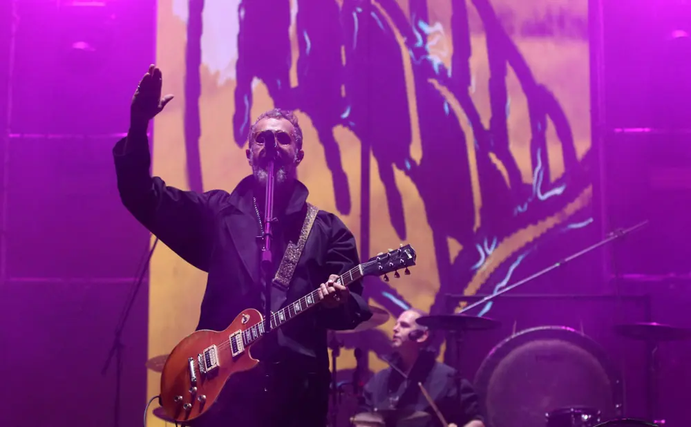 A man with a beard wearing sunglasses and a coat plays a guitar on stage with purple lighting.