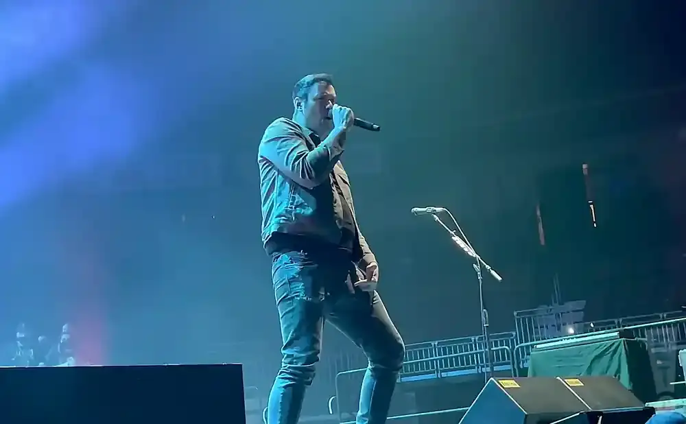A man sings into a microphone on a dimly lit stage with blue spotlights.