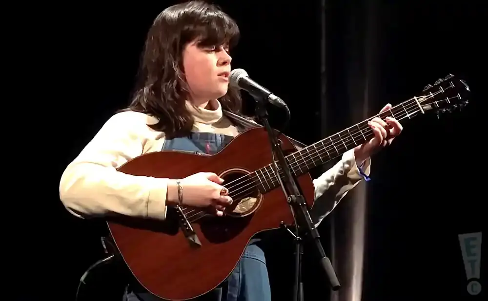 A young person with dark hair sings into a microphone while playing an acoustic guitar on a dark stage.