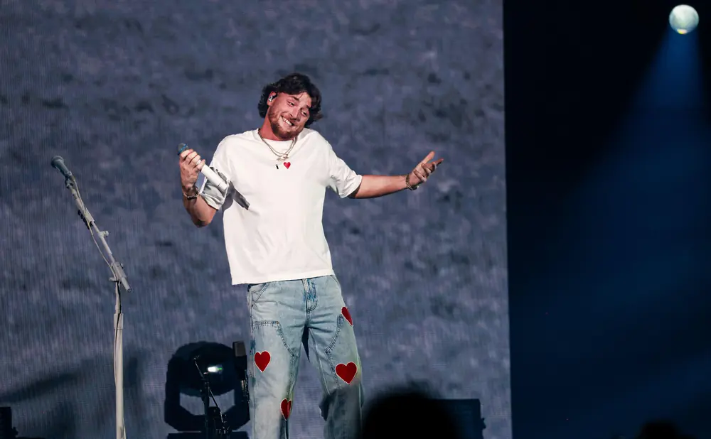 A male performer in jeans with red hearts, white t-shirt, smiles and holds a microphone on stage.