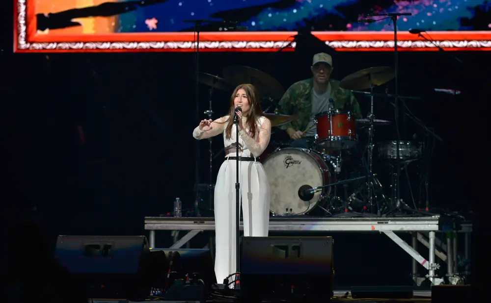 Singer performs on stage in a white outfit with a drummer in the background.