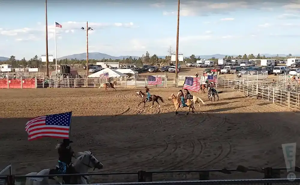 Equestrians ride horses carrying American flags around a rodeo arena, with spectators and vehicles in the background.