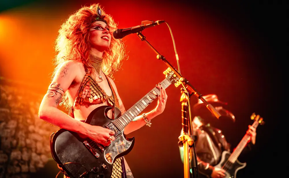 A woman with fiery red hair and dramatic makeup plays a black electric guitar on stage, illuminated by warm, orange stage lights.