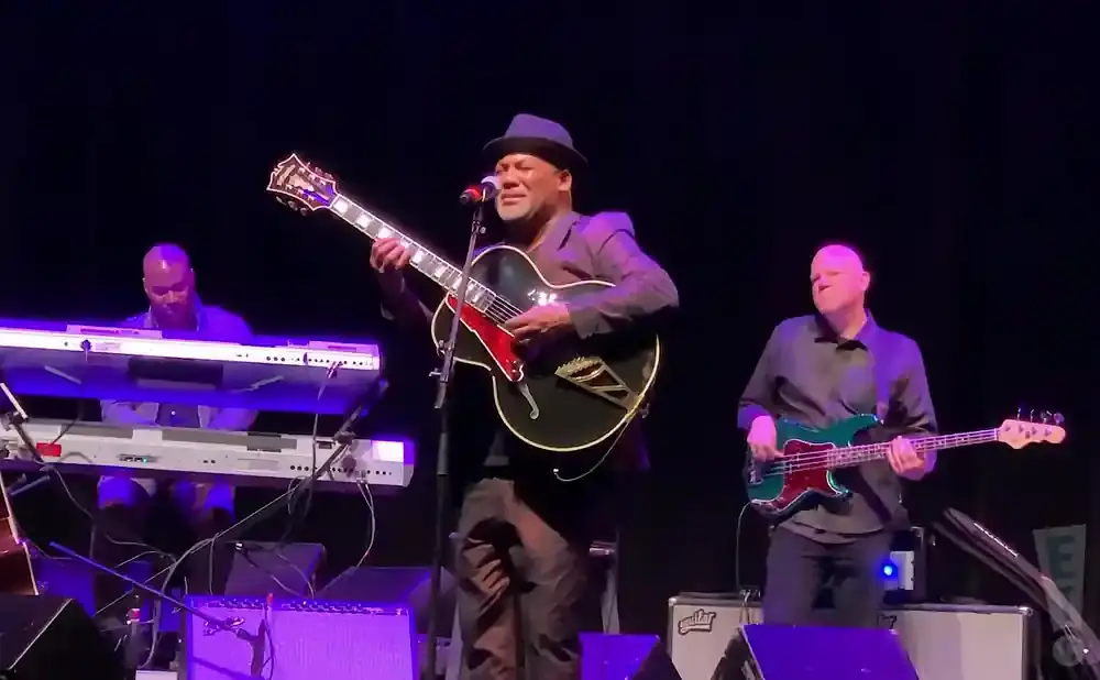 A band performs on stage with a guitarist singing, a keyboardist, and a bassist.