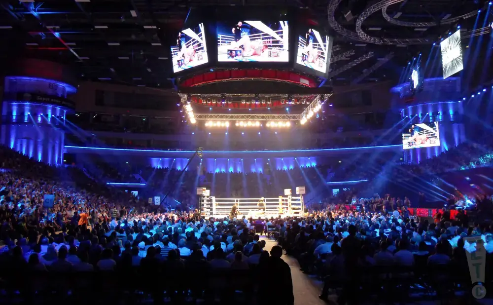 An indoor boxing arena with a brightly lit ring and a large crowd.