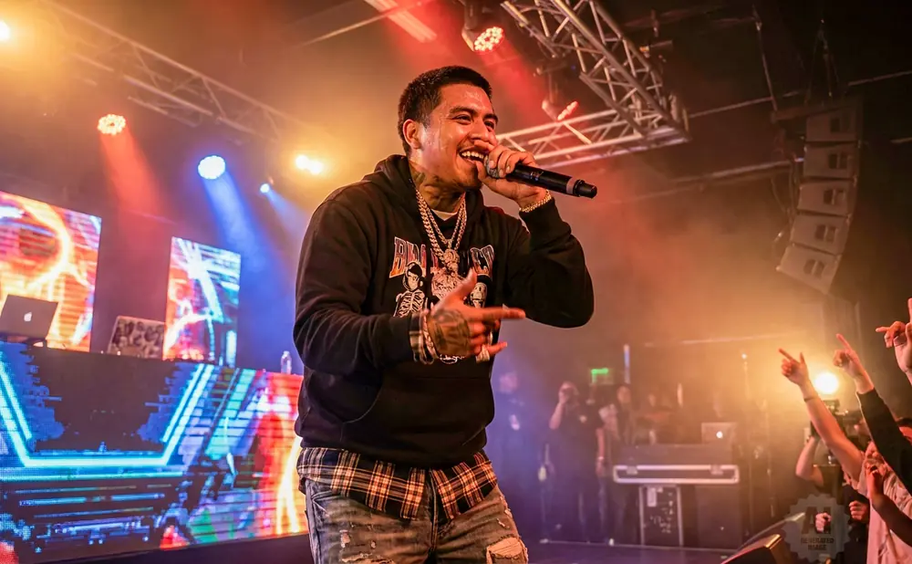 Rapper performs on stage with a microphone, wearing a hoodie, chains, and plaid shirt, with a crowd cheering and DJ setup in the background.