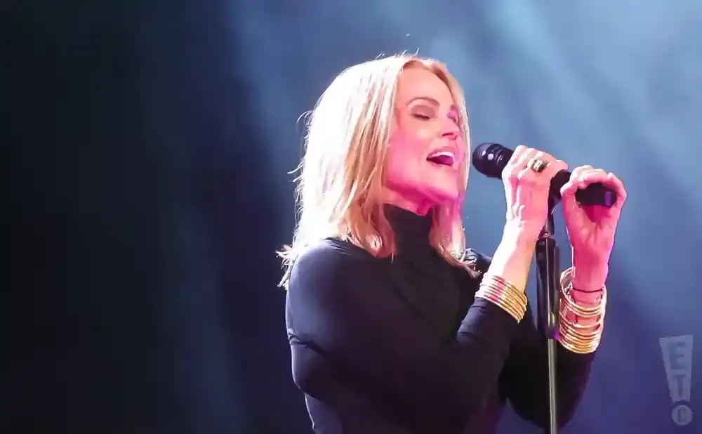Singer with blonde hair, wearing a black turtleneck and gold bangles, holds a microphone and sings.