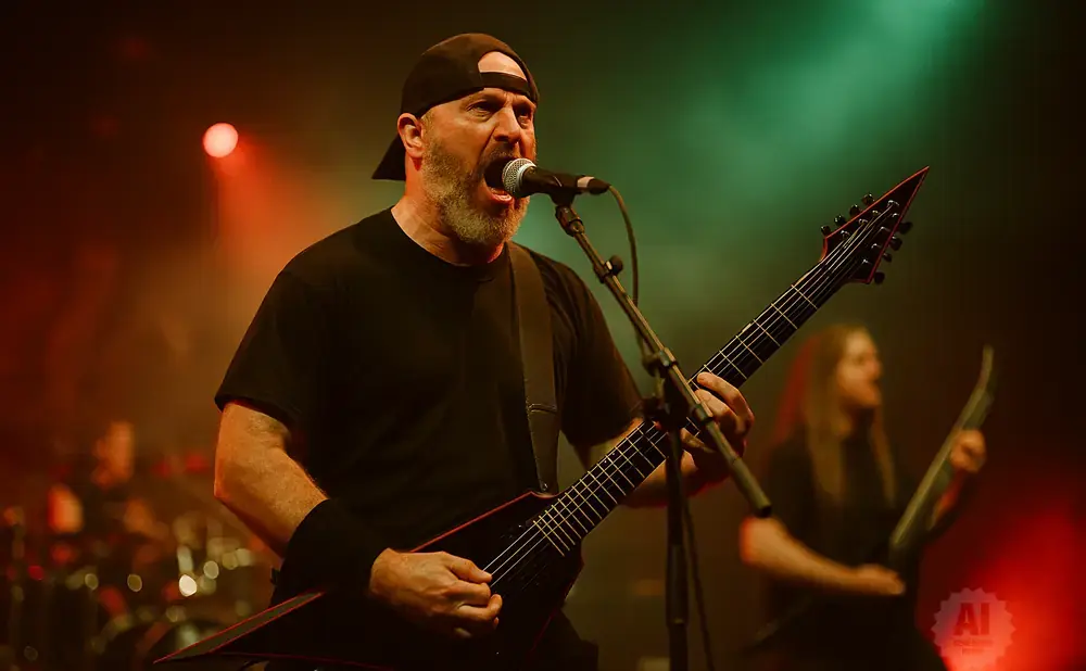 A male heavy metal singer in a black t-shirt and baseball cap sings into a microphone while playing a black V-shaped electric guitar on stage.