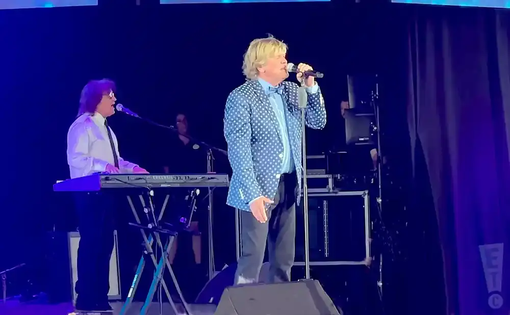 A male singer in a blue polka-dot jacket stands on stage with a microphone, next to another man playing a keyboard.
