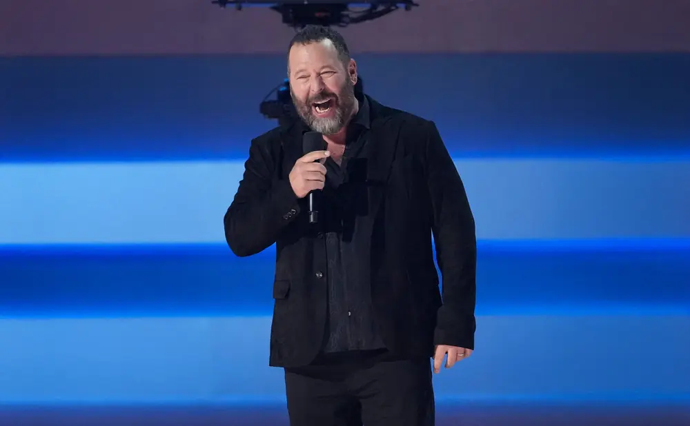 A man with a beard, wearing a black jacket and shirt, holds a microphone and laughs on a blue stage.