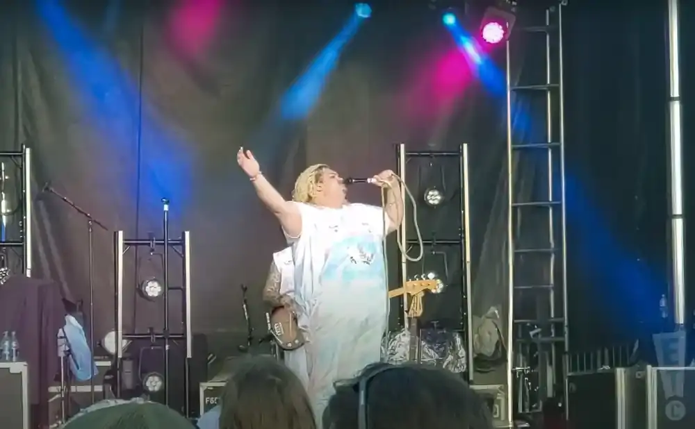 A singer with blonde hair and a white shirt sings into a microphone on a stage with blue and pink lights.