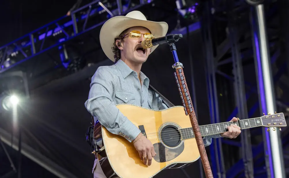 Musician in a cowboy hat and striped shirt plays guitar and sings into a microphone on stage.