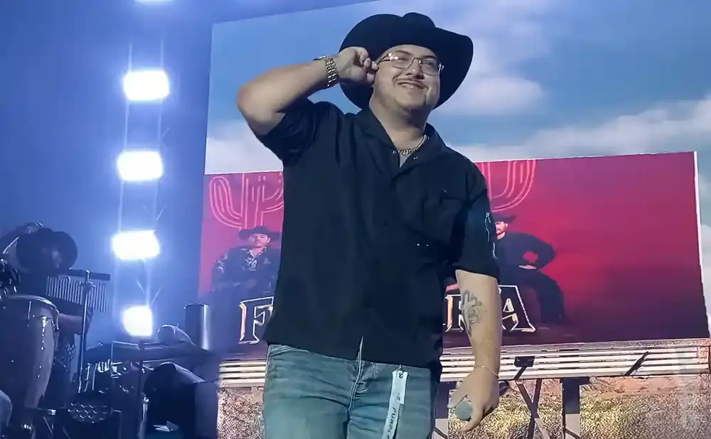 Man in black shirt and cowboy hat salutes on stage with a digital billboard behind him.