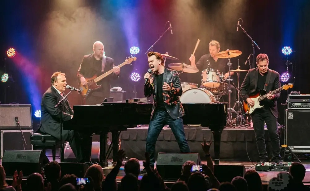 A band performs on stage with a pianist in a suit, a singer in a floral jacket, a guitarist, and a drummer.